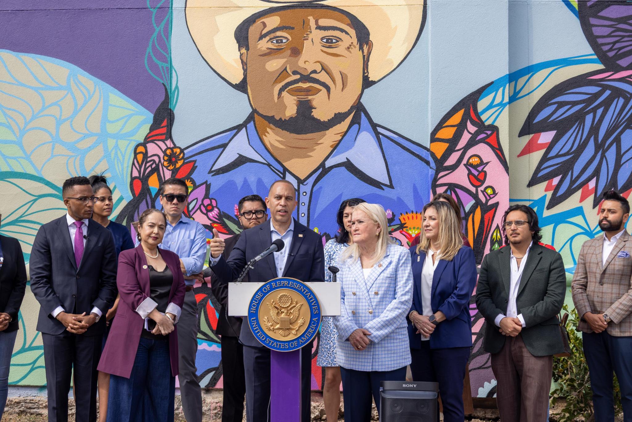 Leader Jeffries speaking from a podium in front of a mural, alongside other press conference participants